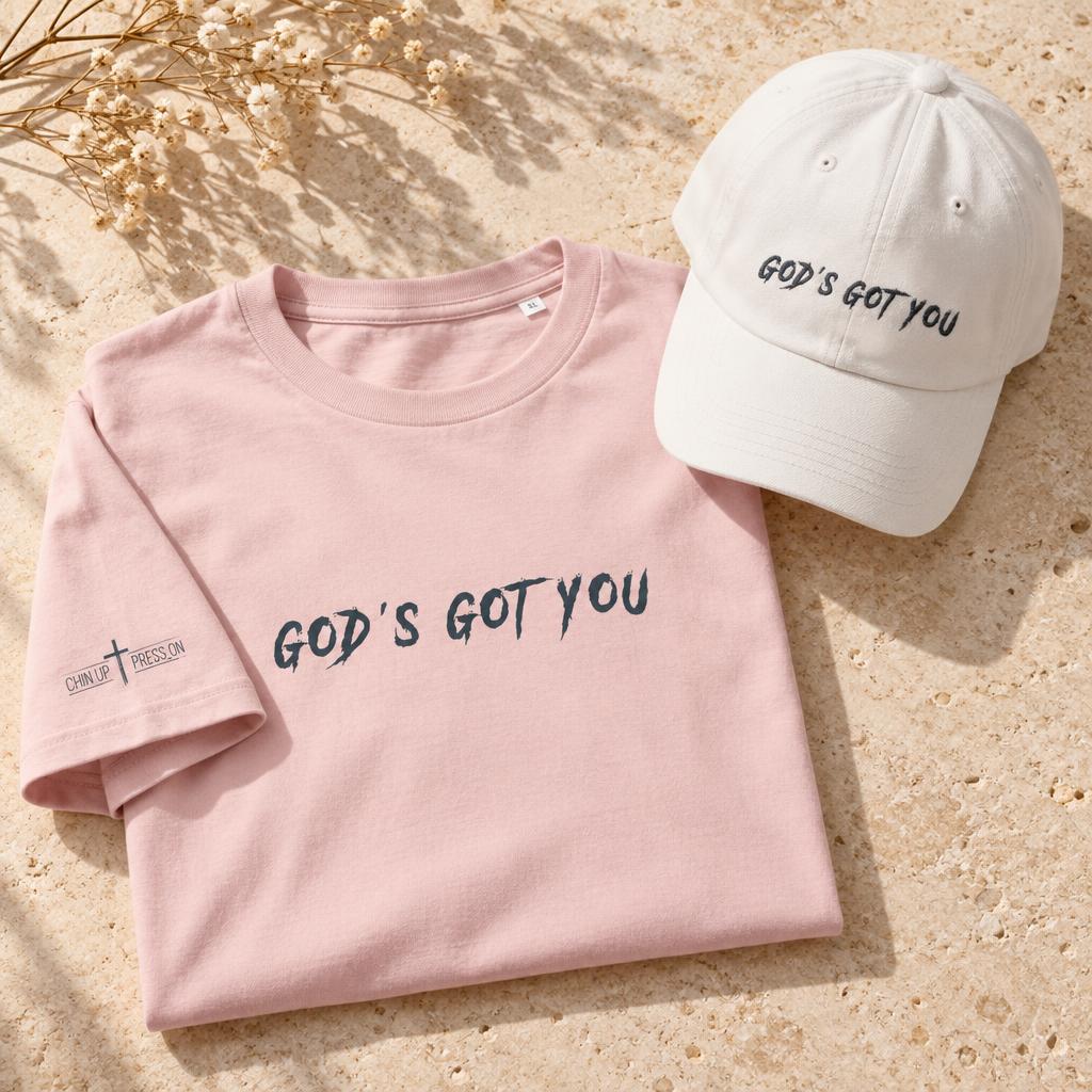 Pink GOD'S GOT YOU tee with CHIN UP ✝ PRESS ON printed on the right sleeve beside a white Dad Hat embroidered with black GOD'S GOT YOU lettering, on warm cream stone.